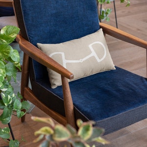 May include: A blue armchair with a wooden frame and a beige pillow with a white horse bit design. The chair is in a room with a green plant in the foreground.