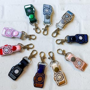 May include: A collection of ten camera-shaped keychains in various colors, each featuring a metal lobster clasp and a snap button, arranged alongside a SanDisk Ultra PLUS 256GB microSDXC UHS-I card in its original retail packaging.