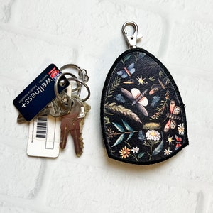 May include: Black leather key chain with a floral and butterfly design. The keychain has a silver metal ring and a silver metal clasp. The text "Wellness" is printed on a blue card attached to a set of keys.