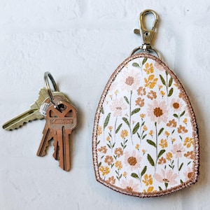 May include: A floral key fob with a metal clasp and a set of keys. The key fob features a white background with a pattern of pink and yellow flowers and green leaves. The fob has a brown border.