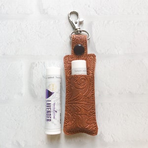 May include: A brown embossed leather lip balm holder with a silver metal lobster clasp and a black snap button, positioned next to a white Young Living Lavender lip balm tube with purple text and a gold seal graphic on the label.