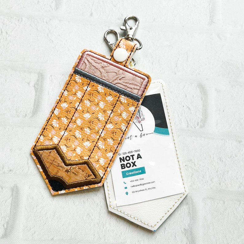 Puede incluir: Un llavero en forma de l&aacute;piz con un patr&oacute;n floral amarillo y blanco, con una etiqueta de cuero blanco adjunta. La etiqueta tiene el texto "NOT A BOX Creations" e informaci&oacute;n de contacto.