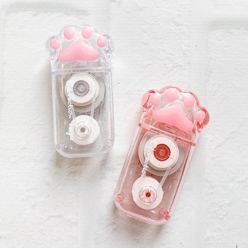 May include: Two clear plastic tape dispensers shaped like a paw print with pink paw pads. The dispensers have a glittery finish and say "Correction Tape" on the side.
