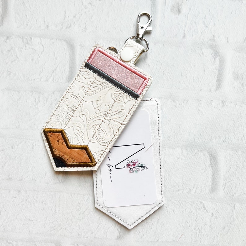 May include: A pencil-shaped keychain pouch featuring a white embossed floral pattern, a pink eraser section, and a cork tip. The pouch includes a silver metal lobster clasp and a white insert card with the letter N and a small floral graphic.