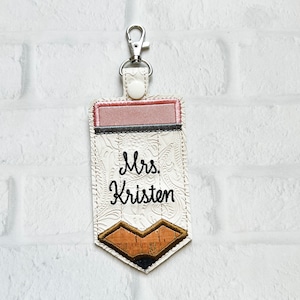 May include: A pencil-shaped keyring featuring a white embossed floral pattern, a pink eraser top, and a cork-textured tip. The text "Mrs. Kristen" is embroidered in black thread across the centre. A silver metal lobster clasp is attached.