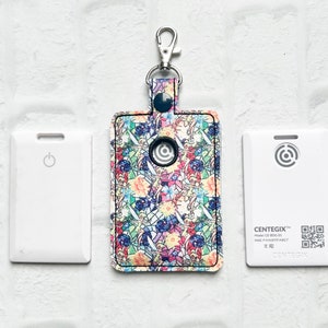 May include: A rectangular key fob holder with a colorful stained-glass design, attached to a silver clip. Two white rectangular devices with a power symbol and a circular design are on either side.