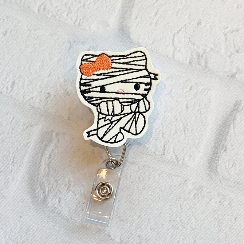 May include: A white mummy costume Hello Kitty character with an orange bow. The character is attached to a clear plastic badge holder with a silver clip.