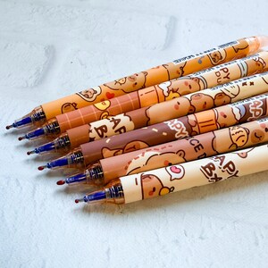 May include: A collection of seven pens with brown and tan bear designs. The pens have blue ink tips and are arranged diagonally. The pen barrels feature various bear illustrations and the text "APY" and "DU".
