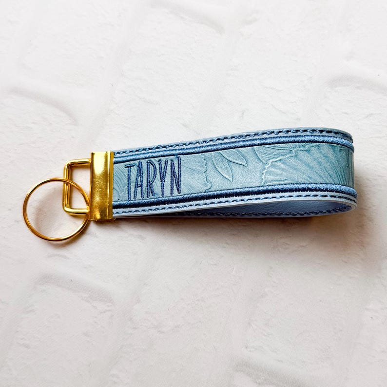 May include: A light blue keychain featuring a textured floral pattern and the name TARYN embroidered in dark blue thread. The strap is secured with a gold-toned metal clasp and includes a matching gold-toned circular ring for keys.