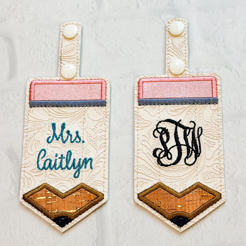 May include: Two rectangular key fobs shaped like pencils with cream-coloured embossed vinyl, pink tops, and brown cork-textured tips. One features the embroidered name Mrs. Caitlyn in teal, while the other displays a black embroidered monogram.