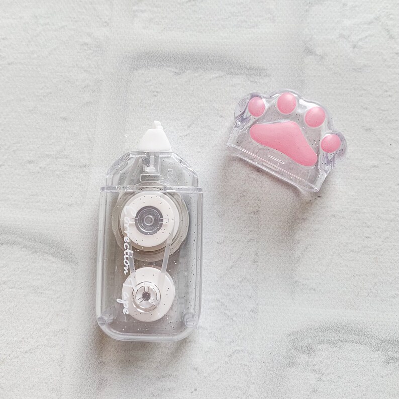 May include: A clear plastic correction tape dispenser with a paw print design. The dispenser has a silver and white tape roll inside. The paw print is pink and has a glitter finish.