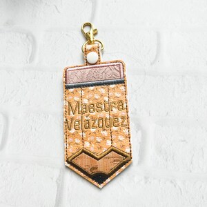 Puede incluir: Un llavero en forma de l&aacute;piz marr&oacute;n y amarillo con un patr&oacute;n floral y el texto "Maestra Velazquez" bordado en hilo dorado.