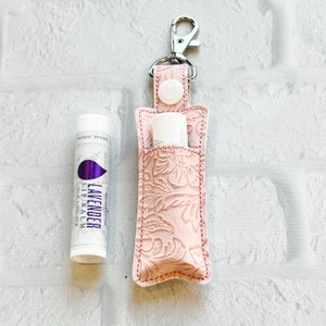 May include: A pink leather lip balm holder with a floral embossed design, a silver-tone clasp, and a white press stud closure. Beside it is a lavender lip balm tube with the text "Lavender" and a purple leaf design.