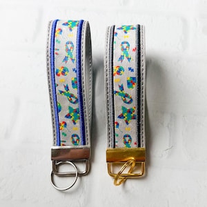 May include: Two wristlet keychains featuring a light gray background with a colorful pattern of puzzle piece ribbons and stars. The left keychain has blue fabric trim and silver hardware, while the right keychain has black trim and gold hardware.
