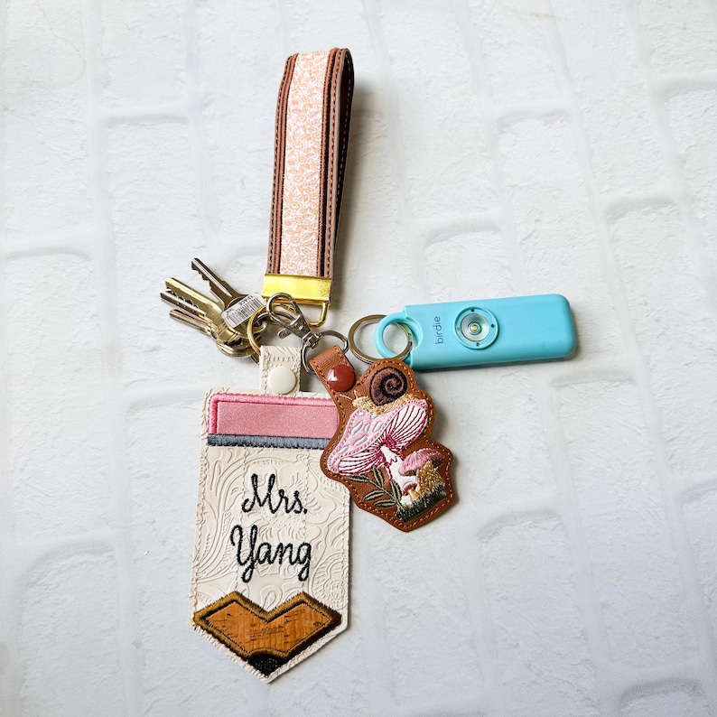 May include: A keychain set featuring a wristlet strap with a peach floral pattern, a pencil-shaped tag embroidered with "Mrs. Yang", a brown leather-look tag with a pink mushroom and snail design, a set of metal keys, and a light blue Birdie alarm.
