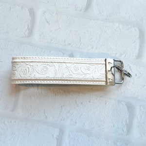 May include: A cream-colored wristlet key fob with a floral embossed design. The key fob has a silver-colored metal clasp and key ring. The wristlet is made of a leather-like material and is set against a white brick wall.