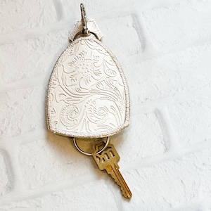 May include: White leather key holder with an embossed floral design. The key holder has a metal ring and a key attached.
