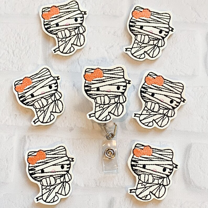 May include: Seven black and white mummy Hello Kitty character iron-on patches with orange bows. The patches are on a white surface. One of the patches has a clear plastic badge holder attached to it.