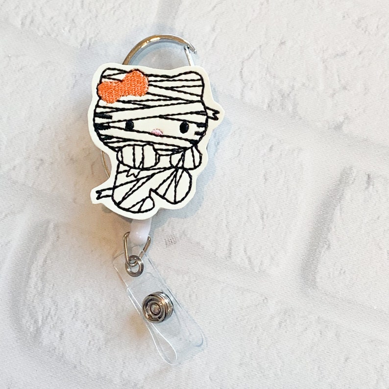 May include: A white and black Halloween themed Hello Kitty character with an orange bow on a retractable badge reel. The character is dressed as a mummy.