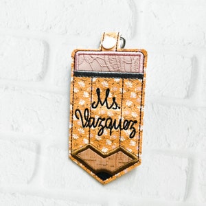 Puede incluir: Un llavero en forma de l&aacute;piz amarillo con un patr&oacute;n floral y el texto "Ms. Vazquez" bordado en &eacute;l.