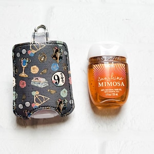 May include: A black and gray fabric hand sanitizer holder with a Harry Potter theme. The holder features images of a golden snitch, a Hogwarts house crest, and the number "9 3/4". The holder is next to a small, clear bottle of hand sanitizer with a yellow and orange label that reads "sunshine mimosa" and "anti-bacterial hand gel with essential oils".