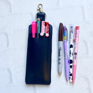 May include: A black leather pen holder with a gold-colored clip, holding several pens and markers. The pens include pink, green, and red Hello Kitty pens, a Sharpie marker, and other colorful pens. The background is a white brick wall.
