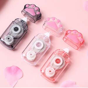 May include: Four clear plastic tape dispensers with paw print designs. Each dispenser has a clear tape roll and a small, clear paw print on the top. The dispensers are on a pink surface.