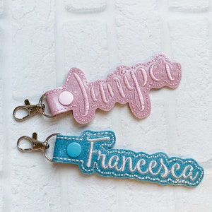 May include: Two personalized keychains featuring glittery fabric with embroidered names. The top keychain is light pink with the name Juniper, and the bottom keychain is bright blue with the name Francesca. Both include silver metal lobster clasps.