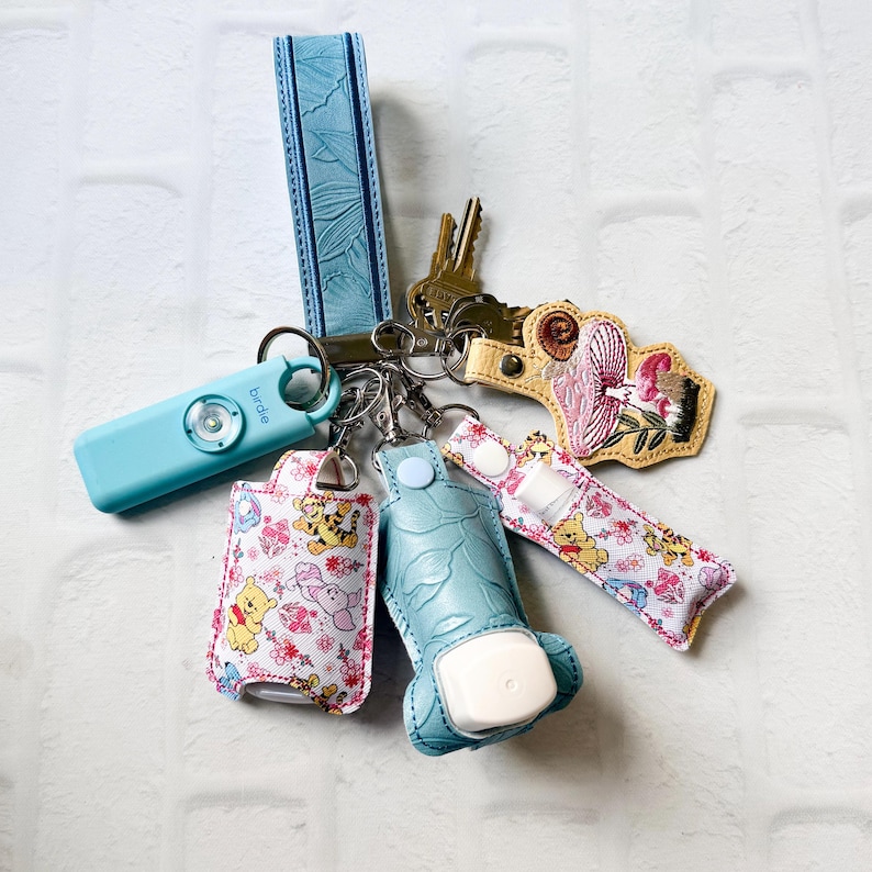 May include: A keychain set featuring a light blue wristlet, a light blue Birdie personal alarm, a yellow embroidered mushroom charm, and two holders with a Winnie the Pooh and Piglet pattern, one holding a small white bottle.