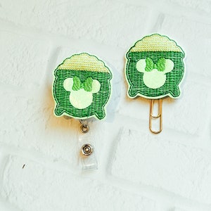 May include: Two green and light green embroidered pot of gold designs. One is a retractable badge holder, and the other is a paper clip. The designs feature a mouse head with a bow. The background is a white brick wall.