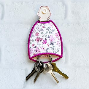 May include: A floral key holder with a white background and a pink floral pattern. The key holder has a white leather top with a snap closure and a pink trim. Several keys hang from the metal ring at the bottom.