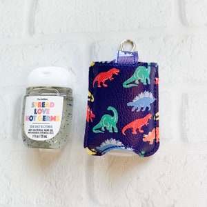 May include: A clear 1 fl oz (29 ml) PocketBac hand sanitizer bottle with a white cap and a label that reads "SPREAD LOVE NOT GERMS". Next to it is a navy blue hand sanitizer holder with a dinosaur pattern in red, green, and blue.