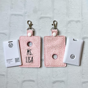 May include: Two pink keychains with a floral design and a clear plastic window. The keychain on the left has the text "MS. LISA" embroidered on it. The keychain on the right has a white card with the text "CENTEGIX" on it.