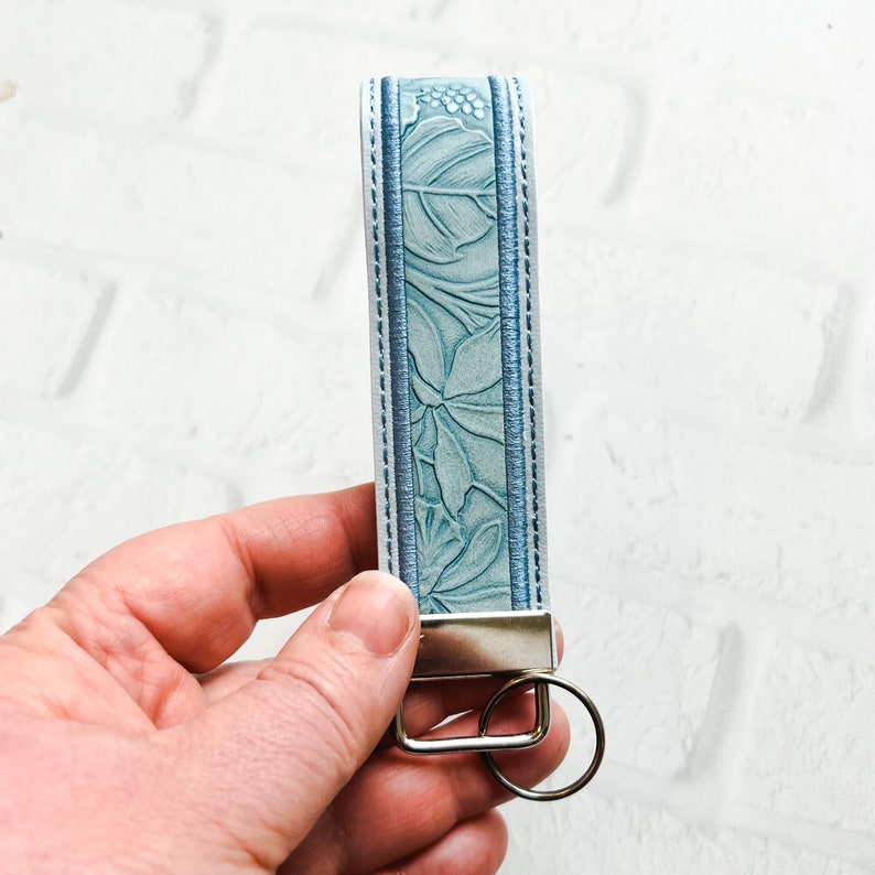 May include: A light blue keychain strap featuring an embossed botanical leaf pattern. The strap is bordered by two thin, darker blue stitched lines and is attached to a silver-toned metal rectangular clamp with a circular split ring attached.