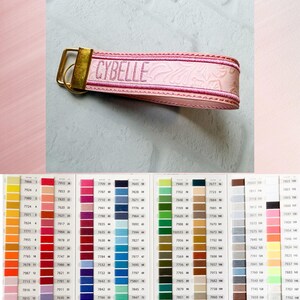 May include: A light pink wristlet keychain featuring an embossed floral pattern and the name CYBELLE embroidered in dark pink thread. The strap is secured with a gold-toned metal clasp and ring, displayed above a color chart with many swatches.