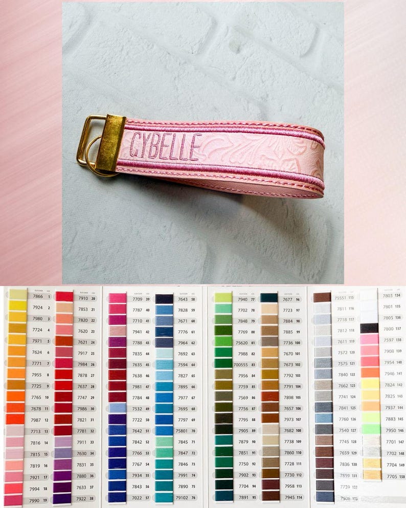 May include: A light pink wristlet keychain featuring an embossed floral pattern and the name CYBELLE embroidered in dark pink thread. The strap is secured with a gold-toned metal clasp and ring, displayed above a color chart with many swatches.