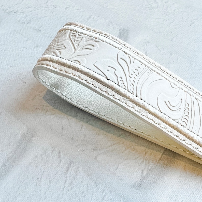 May include: Close-up of a white leather strap with embossed floral and leaf patterns. The strap has a stitched edge and a smooth, rounded end. The design is detailed and the leather appears to be high-quality.