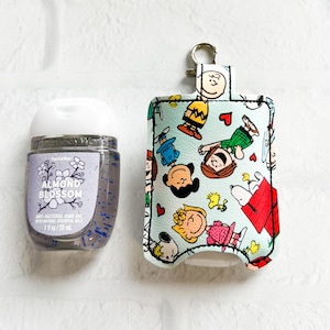 May include: A light blue hand sanitizer holder with a zipper closure and a key ring. The holder is decorated with a Peanuts character print featuring Charlie Brown, Snoopy, and Lucy. The holder is designed to fit a 1 fl oz (29.5 ml) bottle of hand sanitizer.