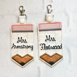 May include: Two rectangular keychains shaped like pencils with silver lobster clasps and white snaps. The top features a pink band, and the bottom has a brown cork-textured tip. The white centre is embossed with a floral pattern and embroidered with the names Mrs. Armstrong and Mrs. Fleetwood in black script.
