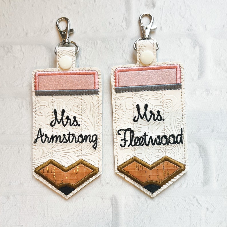 May include: Two rectangular keychains shaped like pencils with silver lobster clasps and white snaps. The top features a pink band, and the bottom has a brown cork-textured tip. The white centre is embossed with a floral pattern and embroidered with the names Mrs. Armstrong and Mrs. Fleetwood in black script.
