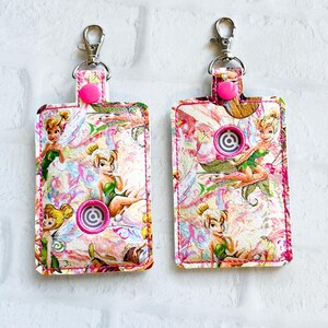 May include: Two keychains with a pink and white floral pattern featuring Tinkerbell illustrations. Each keychain has a silver clasp, a pink snap closure, and a circular design. The edges are stitched with pink thread.