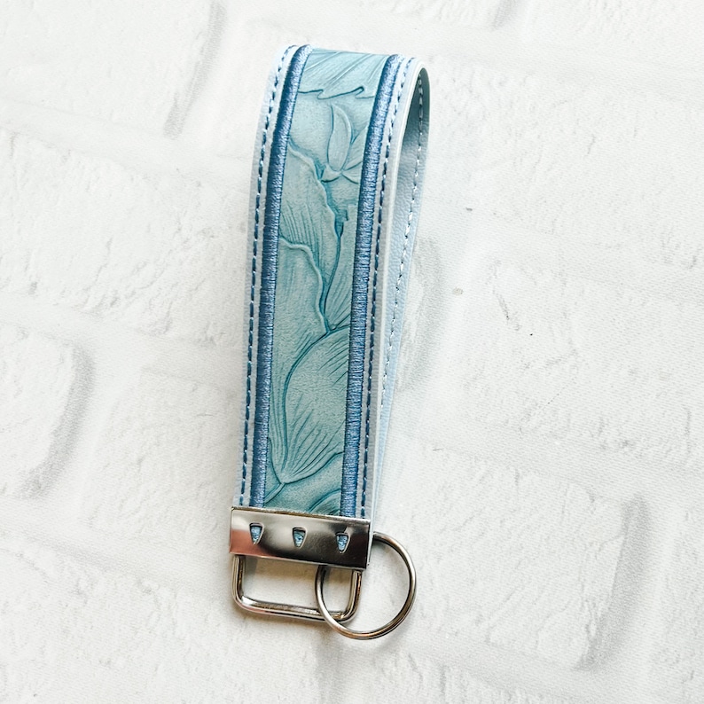 May include: A light blue wristlet keychain featuring a textured floral embossed pattern on the strap. The strap is secured with a silver-toned metal clamp and includes a matching silver-toned split ring for keys, finished with blue stitching.