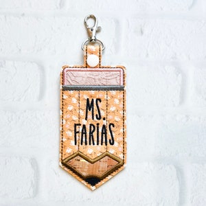 Puede incluir: Un llavero en forma de l&aacute;piz marr&oacute;n y beige con un patr&oacute;n floral y el texto "MS. FARIAS" en negro.