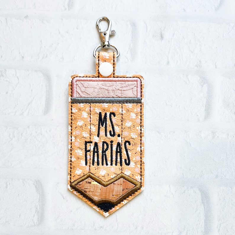 Puede incluir: Un llavero en forma de l&aacute;piz marr&oacute;n y beige con un patr&oacute;n floral y el texto "MS. FARIAS" en negro.