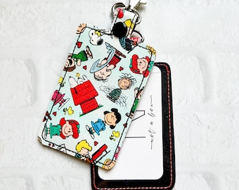 May include: A light blue card holder featuring a cartoon print of Snoopy and friends. The holder has a black border and a silver clasp. The text "not a box" is visible on the card inside.