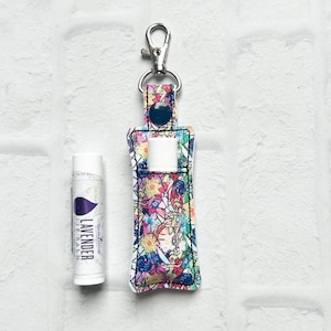 May include: A floral print lip balm holder with a silver clip and a blue snap closure. Next to the holder is a white tube of lavender lip balm. The holder is designed to carry a standard size lip balm.
