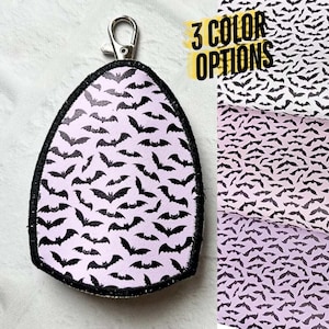 May include: A teardrop-shaped keychain featuring a light purple background with a repeating black bat pattern and a black stitched border. A silver metal lobster clasp is attached to the top. The text 3 COLOR OPTIONS is visible.