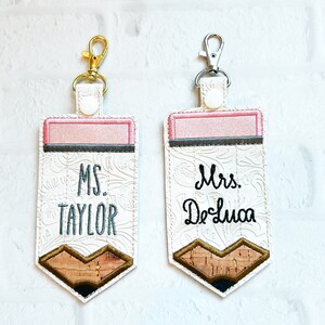 May include: Two rectangular keychains shaped like pencils with white embossed floral patterns. One features a gold-tone clasp and the text MS. TAYLOR, while the other has a silver-tone clasp and the text Mrs. DeLuca. Both have pink erasers.
