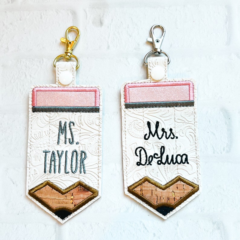 May include: Two rectangular keychains shaped like pencils with white embossed floral patterns. One features a gold-tone clasp and the text MS. TAYLOR, while the other has a silver-tone clasp and the text Mrs. DeLuca. Both have pink erasers.