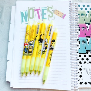 May include: Six yellow pens with Pikachu designs are arranged on a lined notebook page. The pens have various Pikachu illustrations and Japanese text. A notebook with the word "NOTES" and a pencil sticker is in the background.